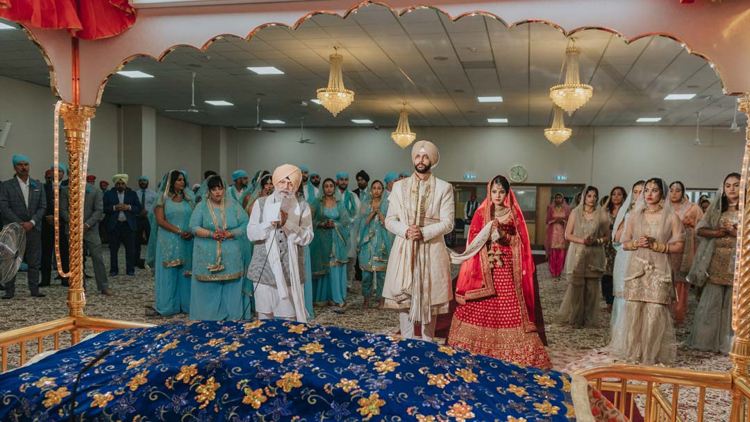 A Sikh Bride and Groom stand with the whole congregation to join in the Ardass at Gurdwara Hall.