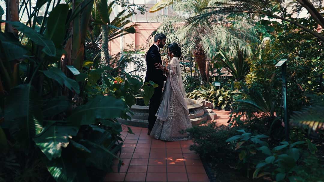 The couple is dressed in Asian Wedding attire, as they stand in the Subtropical Glasshouse, Birmingham Botanical Gardens for their Asian Wedding Photography. They are surrounded by lush greenery, including palm trees and other plants. 
