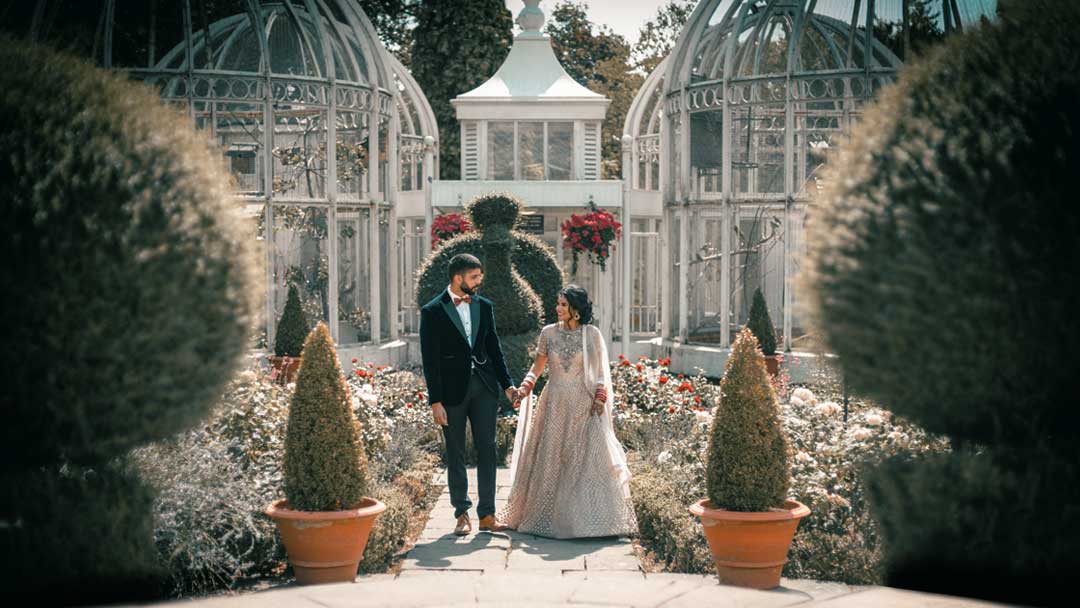 The couple is dressed in Asian Wedding attire, as they stand in the Subtropical Glasshouse, Birmingham Botanical Gardens for their Asian Wedding Photography. They are surrounded by lush greenery, including palm trees and other plants. 