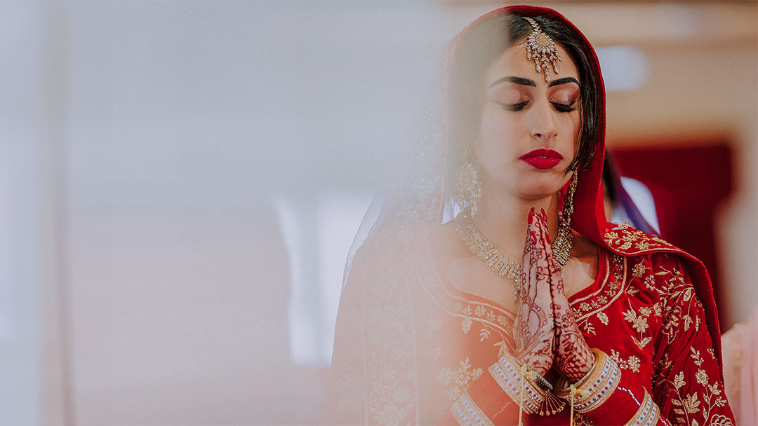 A Sikh bride in traditional Punjabi red attire, hands folded in worship at Sri Guru Singh Sabha Gurdwara Kettering.