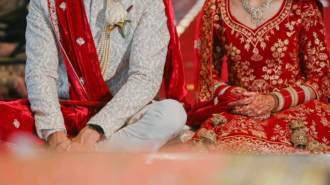 At Kettering Sikh Temple, a bride holds her husband's palla with wearing intricate henna designs, red bangles, and gold jewellery.