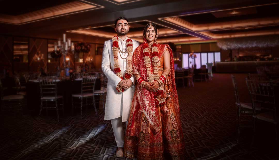 An Indian couple stands together in a wedding hall in Wolverhampton, their traditional Indian attire vibrant against the backdrop of elegant chandeliers and tables set for the Indian wedding reception.