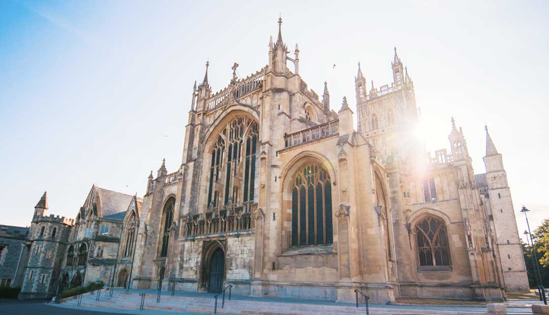 Gloucester Cathedral, a gothic church with towers and spires, is illuminated, creating a lens flare effect. The cathedral, with its ancient and solemn stone facade, perfect for a wedding photoshoot.