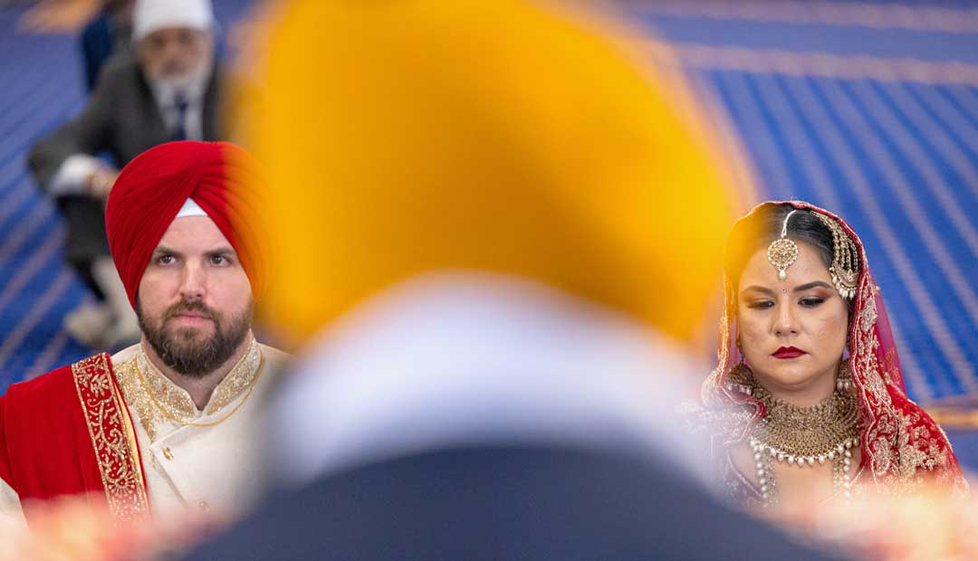 A couple in traditional Punjabi attire sits in front of Sri Guru Granth Sahib Ji during the Anand Karaj ceremony at the Leamington Spa Gurdwara.