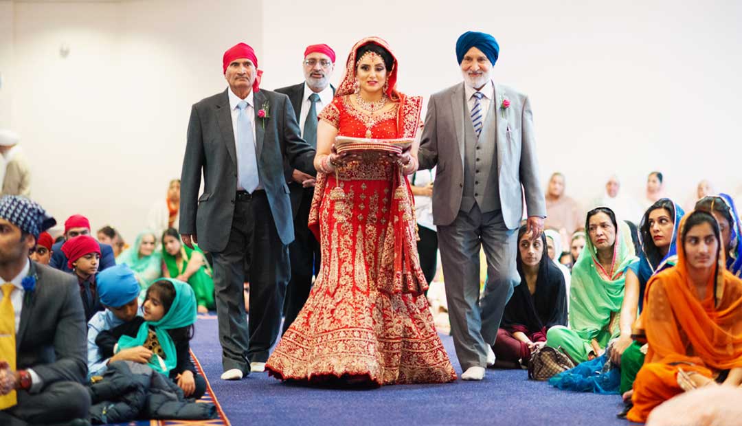 A Sikh bride in a traditional red outfit walks down the aisle at the Leamington Sikh temple, accompanied by two men in turbans.