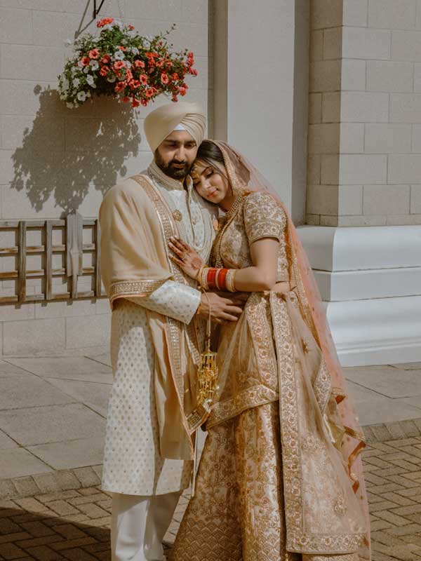 couple poses for photo Leamington Gurdwara A newlywed couple poses for a photo, the bride's head resting on her husband's chest. They stand against a backdrop of Leamington Spa Gurdwara with a flower basket hanging from its side.