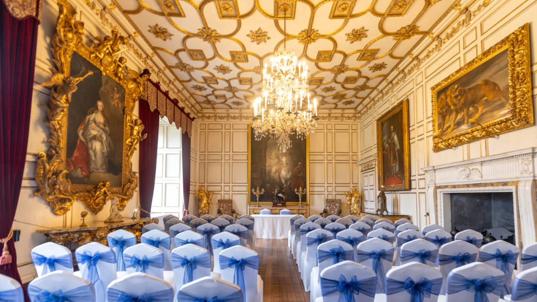 An elegant wedding ceremony setup in a historic State Dining Room Warwick Castle. The room features ornate wood panelling, a high ceiling with intricate gold details, and a large painting on the wall. 