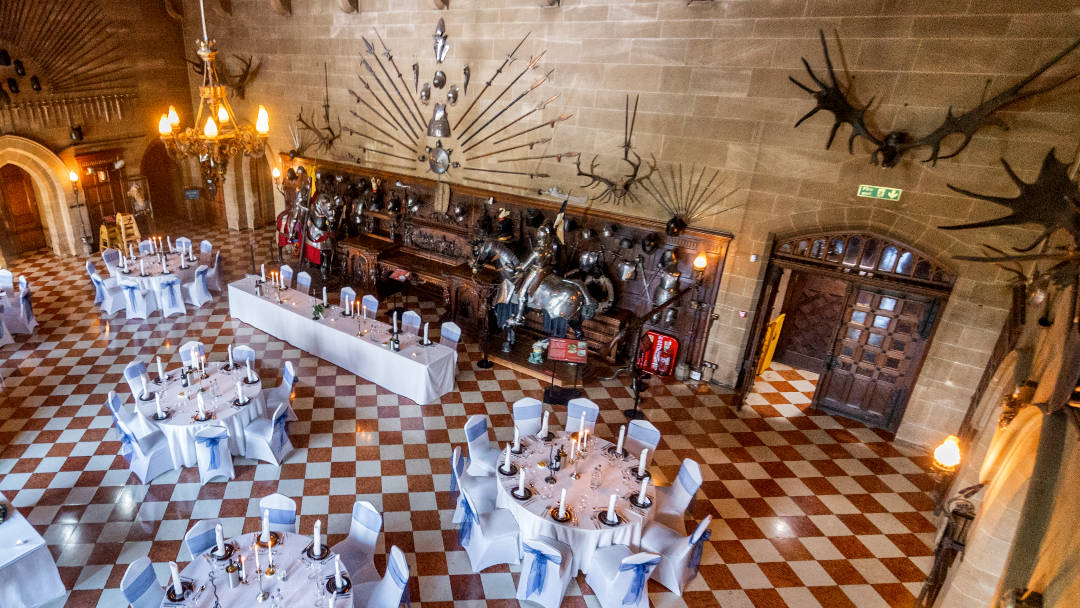 An elegant wedding ceremony setup in a historic Great Hall Warwick Castle. The room features ornate wood panelling, a high ceiling with intricate gold details, and a large painting on the wall. 