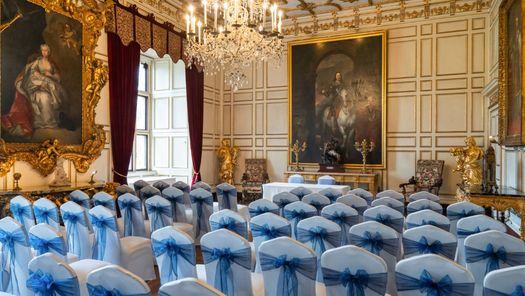 The Warwick Castle State Dining Room photo features rows of white chairs with blue sashes facing a small table with a white tablecloth. A stunning crystal chandelier adorns the high ceiling.